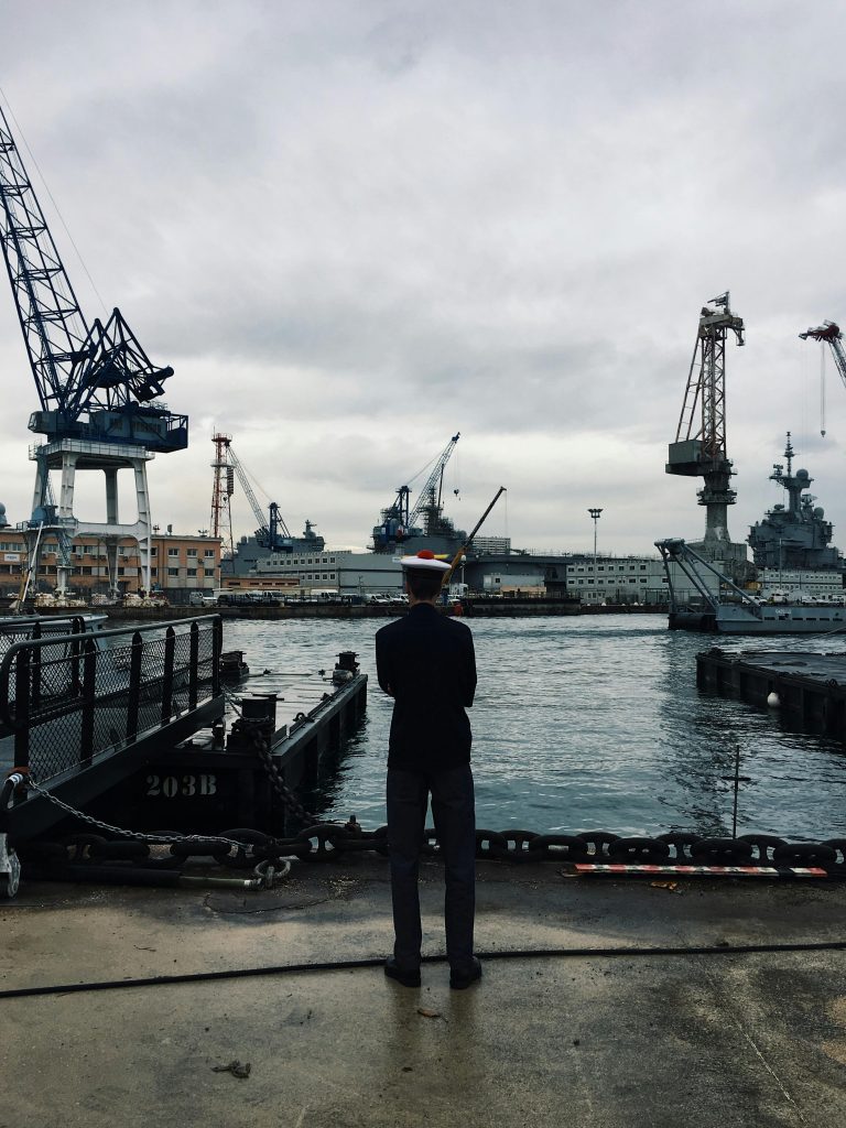 Navy officer oversees ships at Toulon's industrial harbor under a cloudy sky.